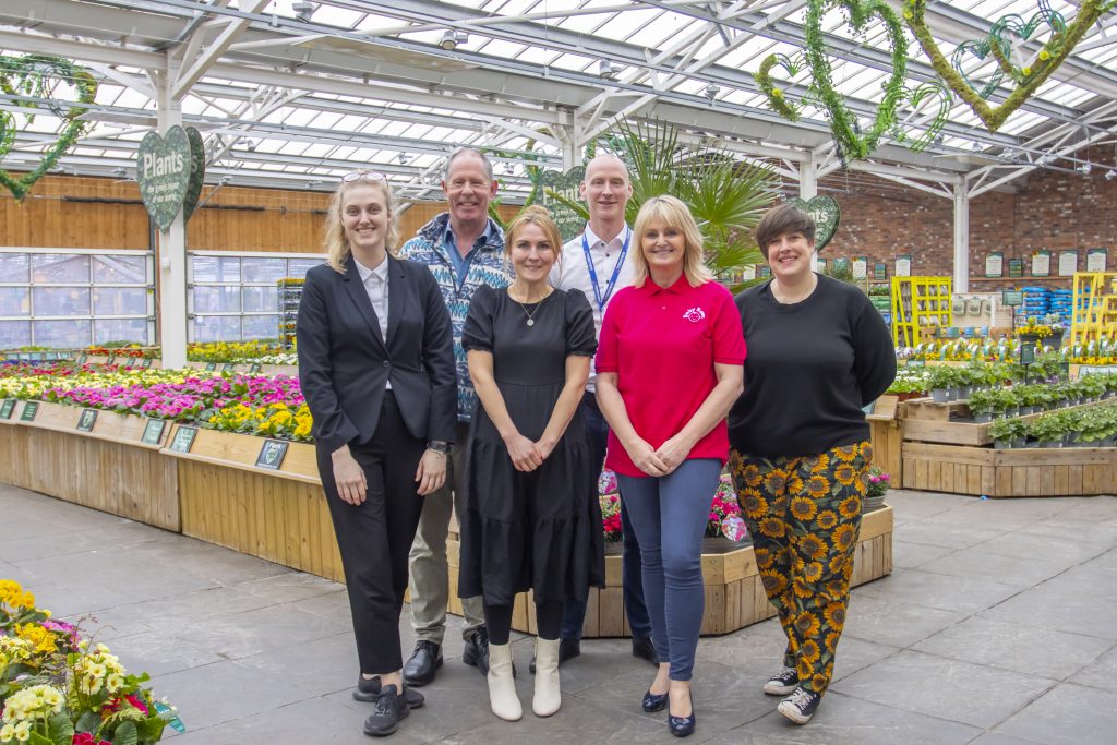 Image of 6 people smiling in a greenhouse with plants and foliage in the background.