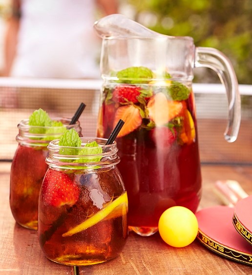 Image of two glasses and a jug of Pimms cocktail with lemon, strawberries, mint and cucumber garnish and black straws.